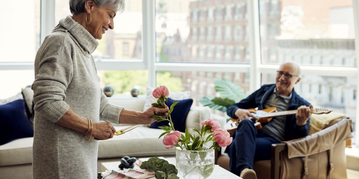 Senior living residents in their apartment, older woman arranging flowers at counter while older man plays guitar in background in chair.