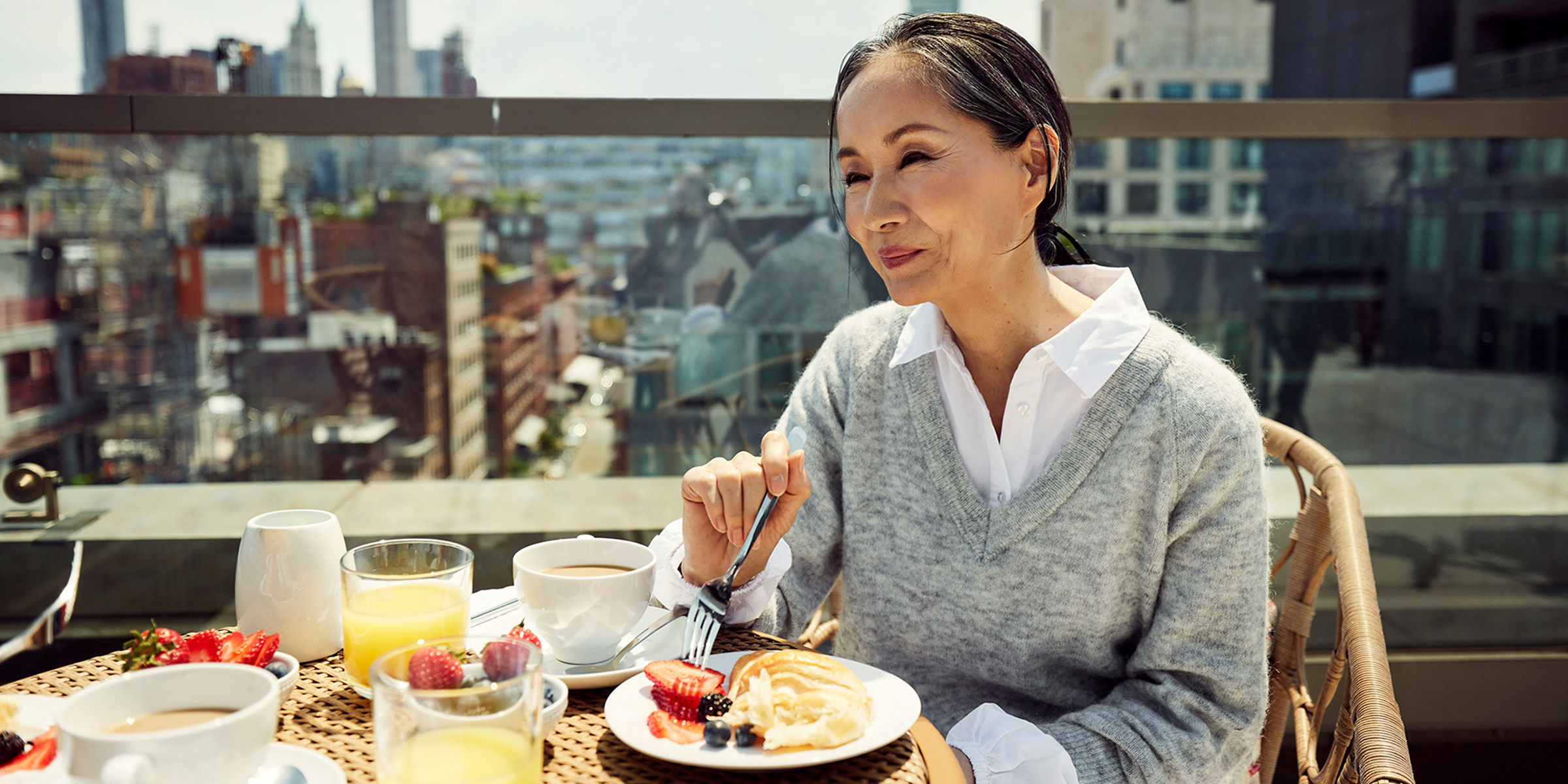 Older woman eating breakfast on the rooftop with the city skyline splayed out behind her